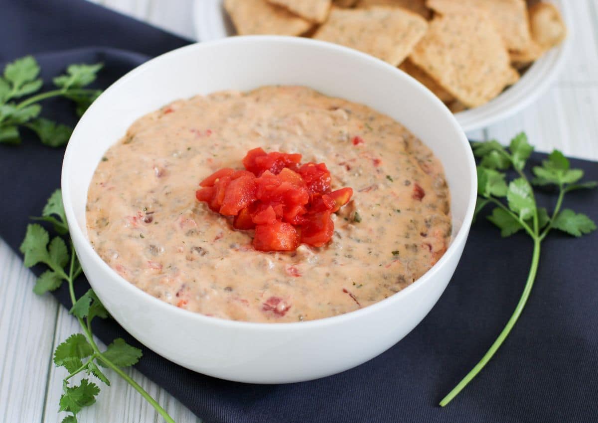 A bowl of creamy cheese dip topped with diced tomatoes, placed on a dark napkin with fresh cilantro and a plate of chips in the background.