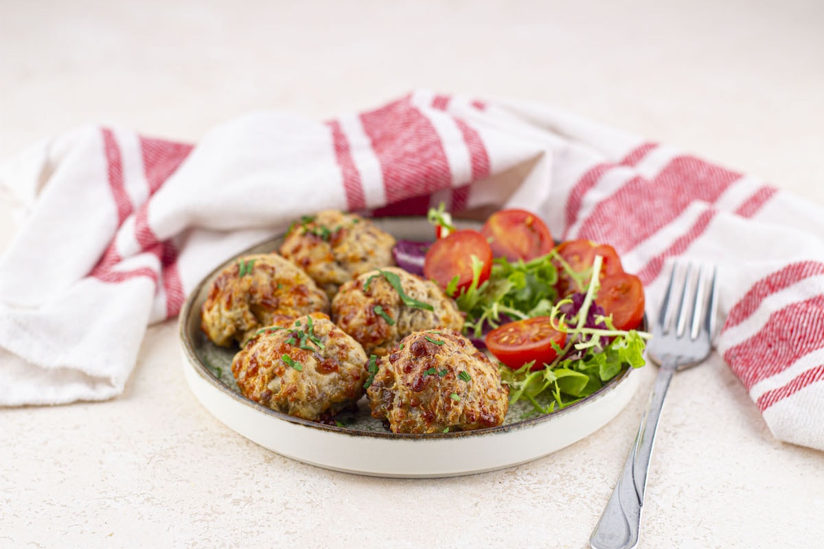 A plate with five meatballs, a side of cherry tomato salad, and leafy greens; a fork is beside the plate and a red-striped towel is in the background.