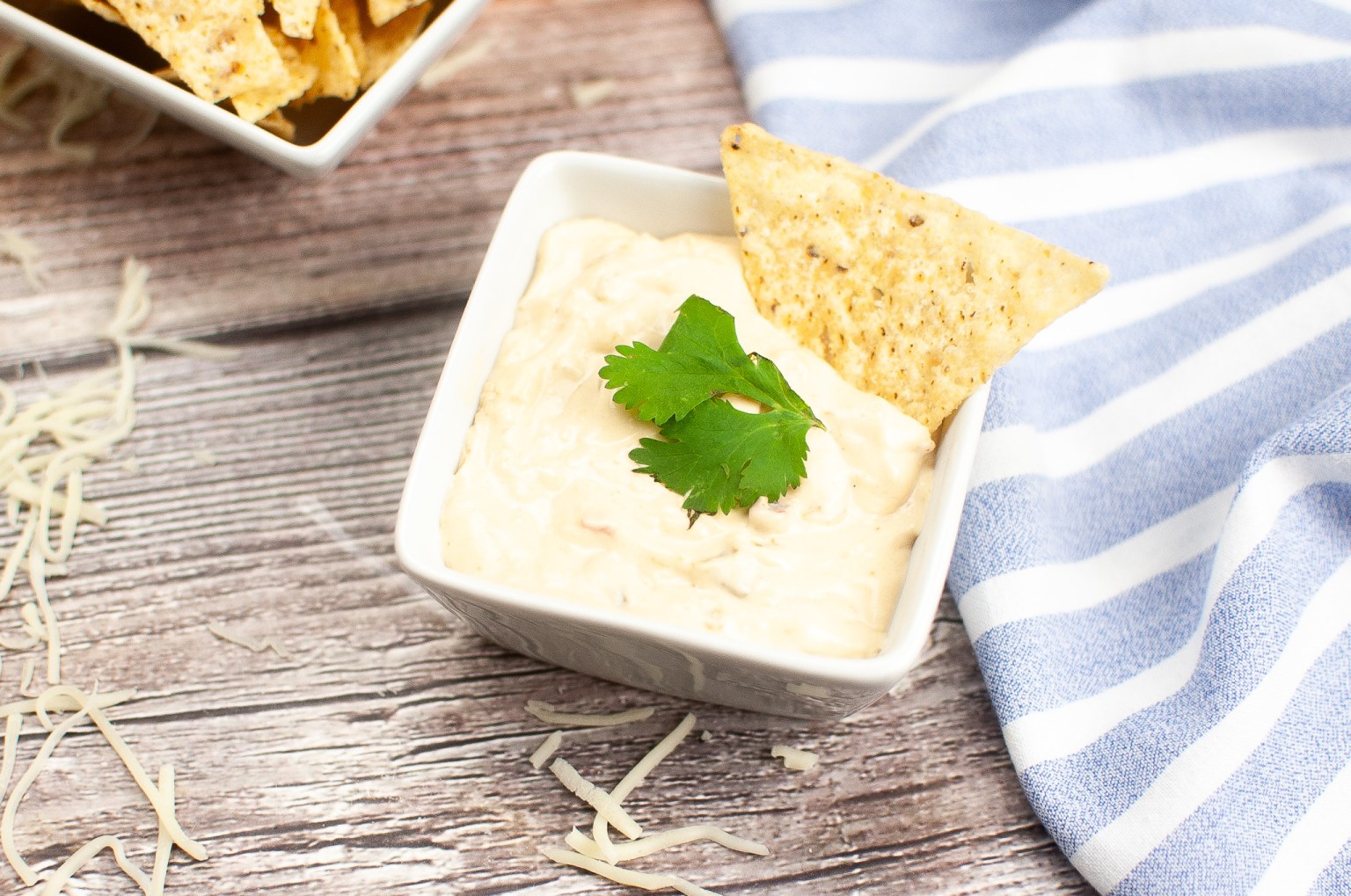 A square bowl of creamy cheese dip garnished with cilantro and a tortilla chip, placed on a wooden surface near a striped blue towel and shredded cheese.