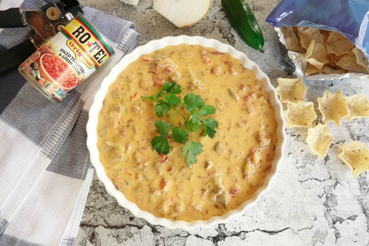 A bowl of cheesy dip with tomatoes and green chiles, garnished with cilantro, sits next to tortilla chips, a can of Ro-Tel, and a kitchen towel.