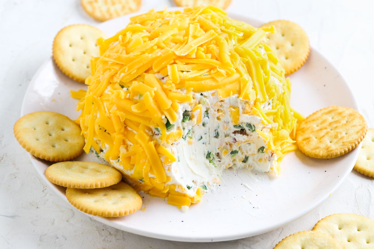 A cheese ball covered in shredded cheddar with herbs inside, served on a white plate surrounded by round crackers. A section of the cheese ball has been scooped out.