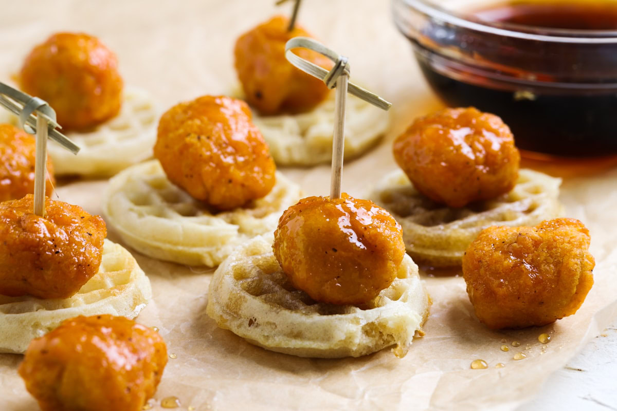 Mini waffles topped with saucy chicken bites, secured with toothpicks, on parchment paper with a bowl of syrup in the background.