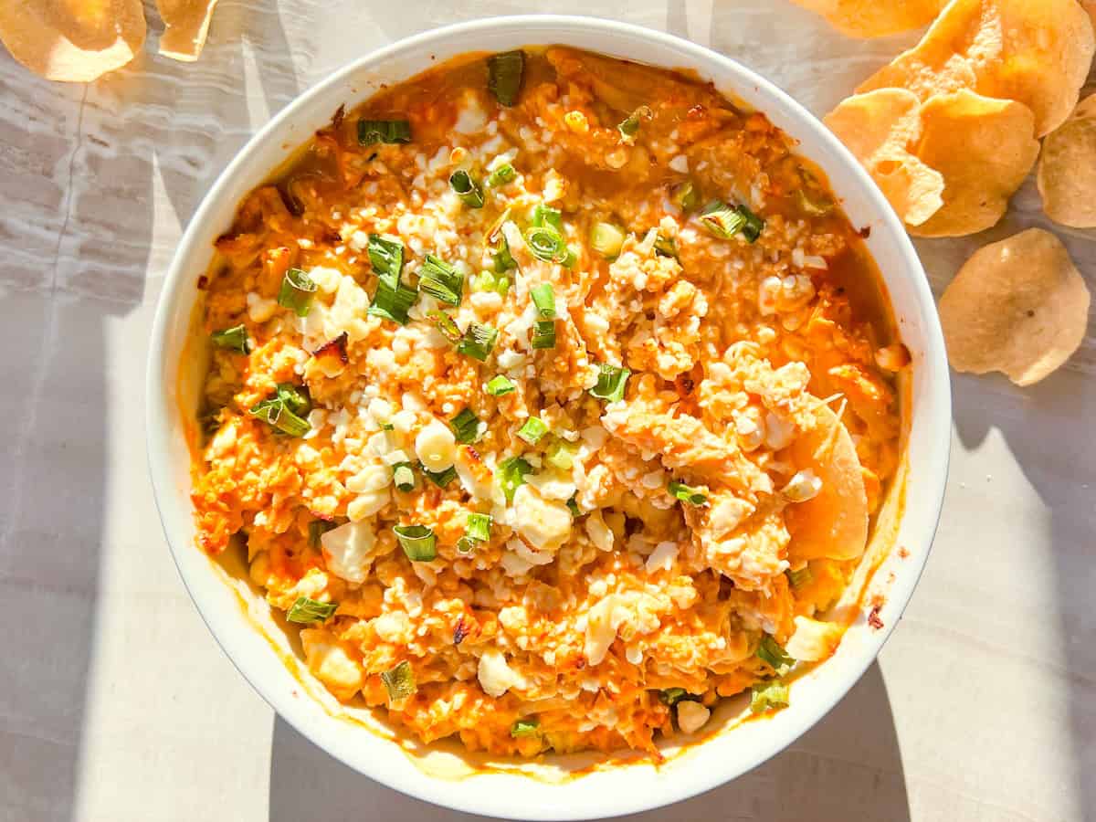 A bowl of mixed cheese dip topped with chopped green onions and seasonings, with round chips on the side.
