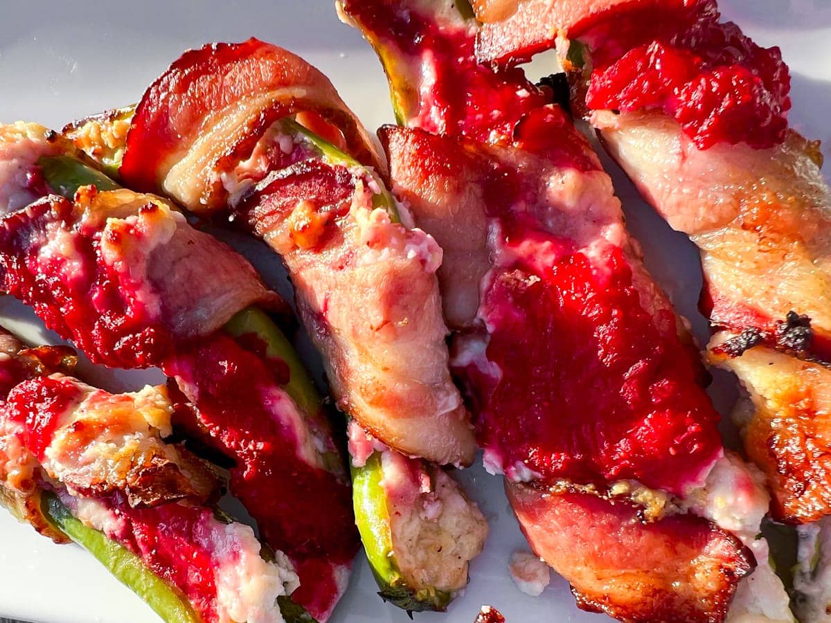 Bacon-wrapped jalapeรฑo poppers stuffed with cheese and topped with bright red raspberry sauce, arranged on a white plate.
