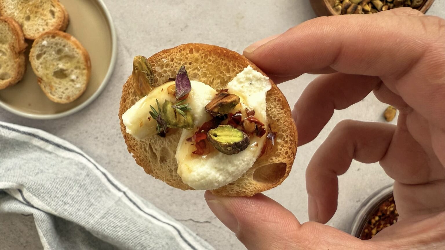 A hand holds a crostini topped with ricotta cheese, honey, pistachios, herbs, and chili flakes. Slices of bread and a bowl of pistachios are visible in the background.