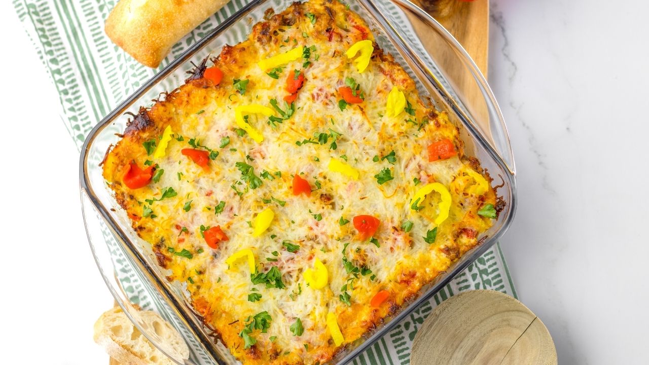 A baked casserole topped with melted cheese, chopped parsley, and sliced yellow and red peppers in a glass dish, placed on a striped cloth with bread beside it.