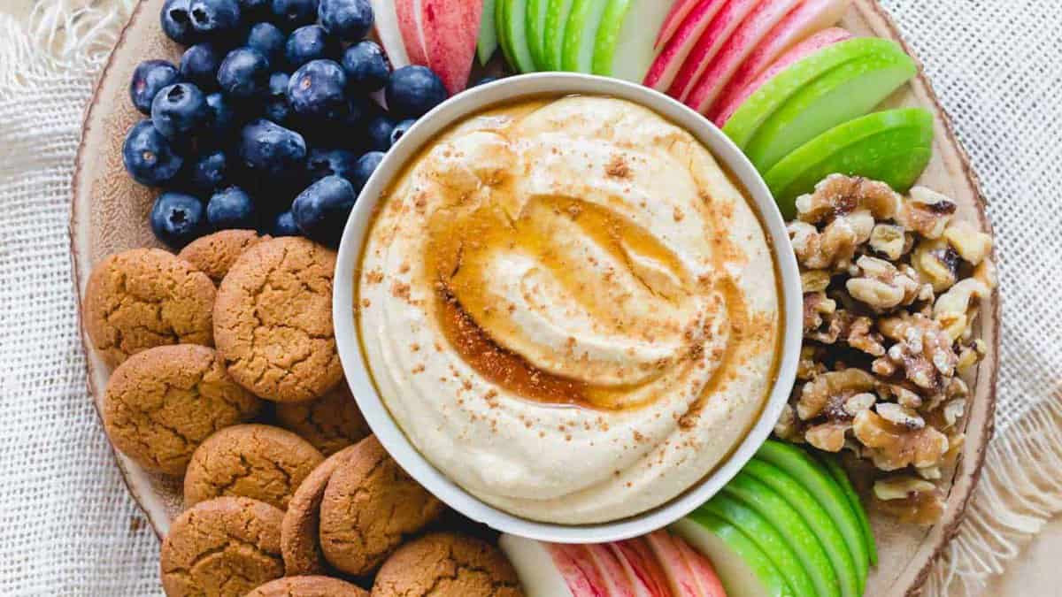 A plate with blueberries, gingersnap cookies, green and red apple slices, walnut pieces, and a bowl of dip topped with honey and cinnamon in the center.