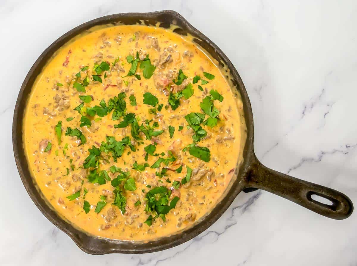 A cast-iron skillet filled with creamy cheese dip containing ground meat and topped with chopped fresh cilantro, on a white marble surface.
