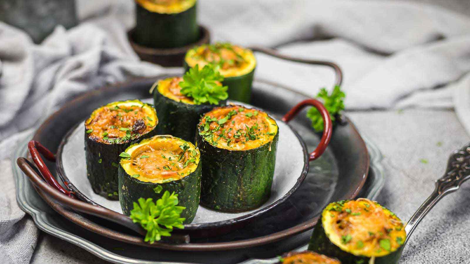 Slices of zucchini stuffed with a baked filling, garnished with chopped herbs and parsley, arranged on a plate with a fork and napkin in the background.