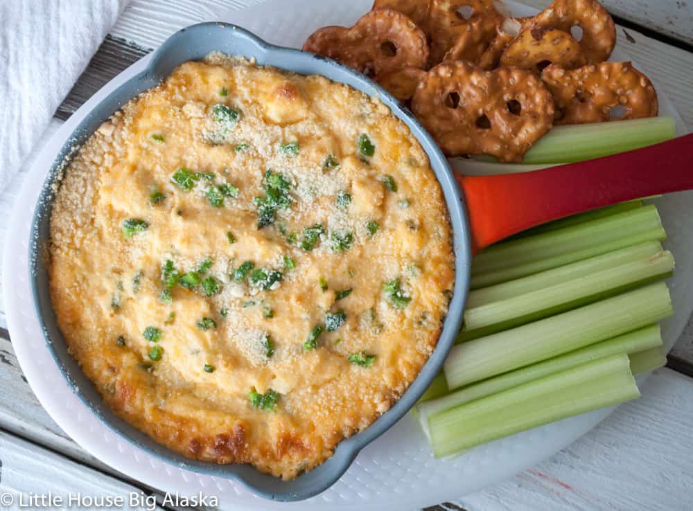 Baked dip garnished with herbs in a skillet, served with celery sticks and pretzel crisps on a white plate.