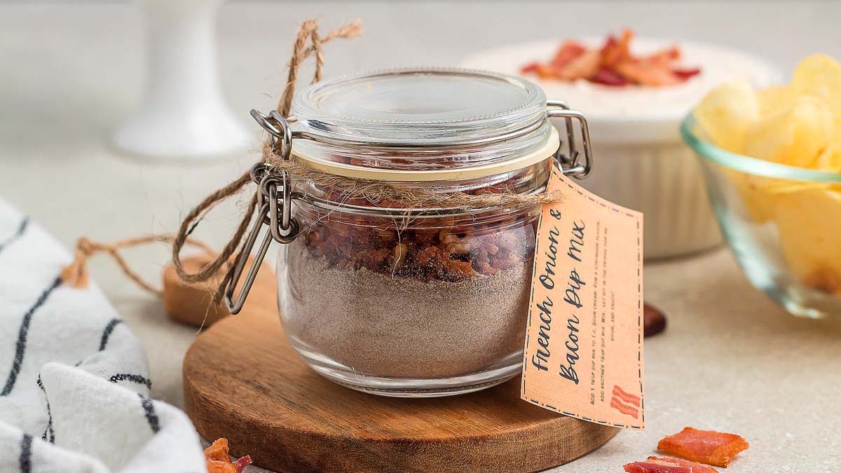 A glass jar filled with French onion and bacon dip mix sits on a wooden board, with a handwritten label attached. Other snacks and chips are visible in the background.