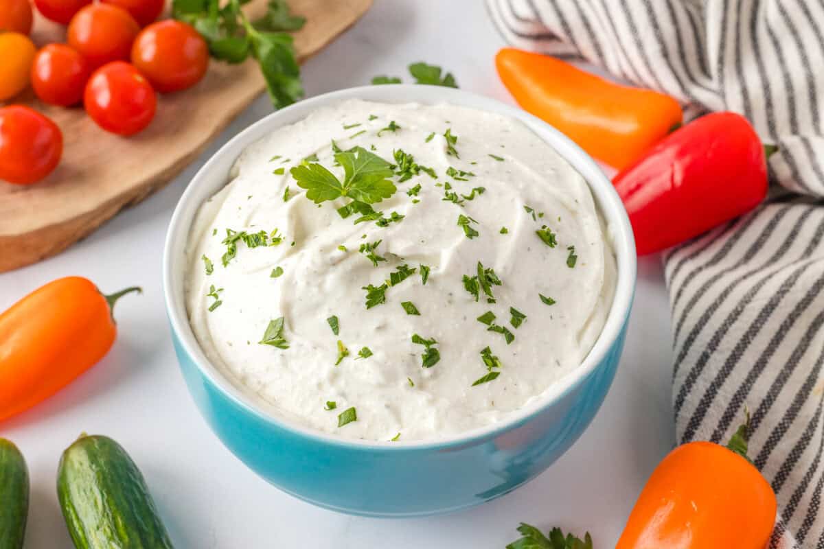 A blue bowl filled with creamy white dip garnished with chopped herbs, surrounded by cherry tomatoes, mini bell peppers, and cucumbers.
