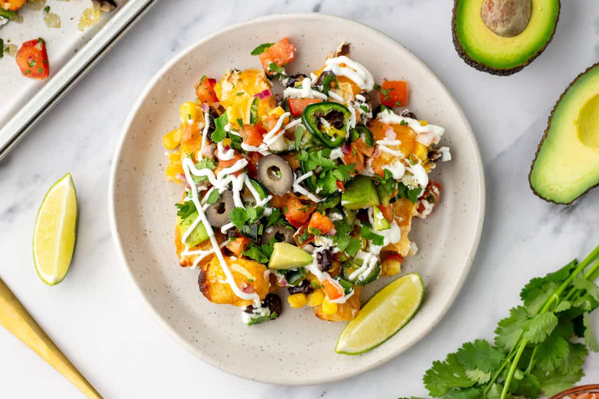 A plate of loaded nachos topped with cheese, black olives, jalapenos, chopped tomatoes, avocado, cilantro, and drizzled with white sauce, with lime wedges on the side.