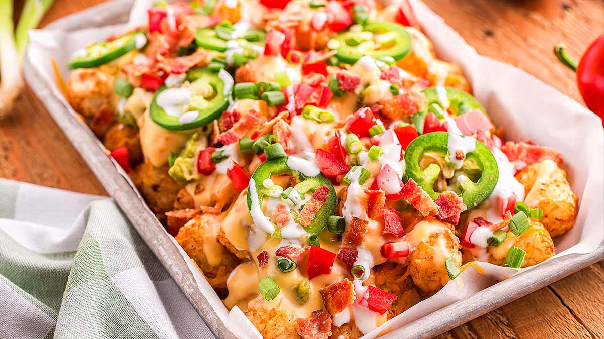 A tray of loaded tater tots topped with melted cheese, bacon, diced tomatoes, sliced jalapeรฑos, green onions, and a drizzle of creamy sauce.