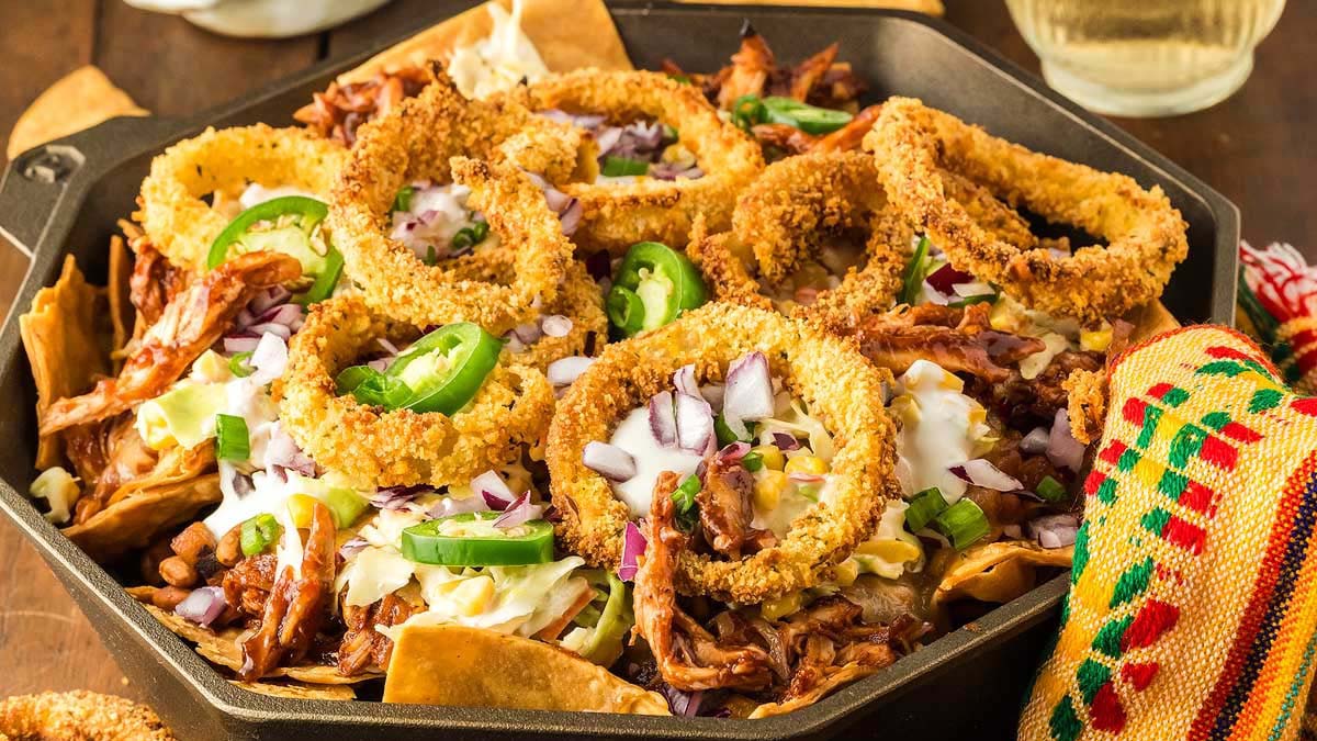 A skillet filled with nachos topped with shredded meat, fried onion rings, sliced jalapeรฑos, red onions, green onions, and a dollop of sour cream.