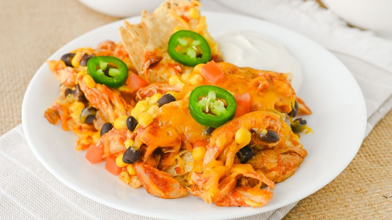 A plate of chicken nachos topped with melted cheese, black beans, corn, diced tomatoes, sliced jalapeรฑos, and served with tortilla chips and a side of sour cream.