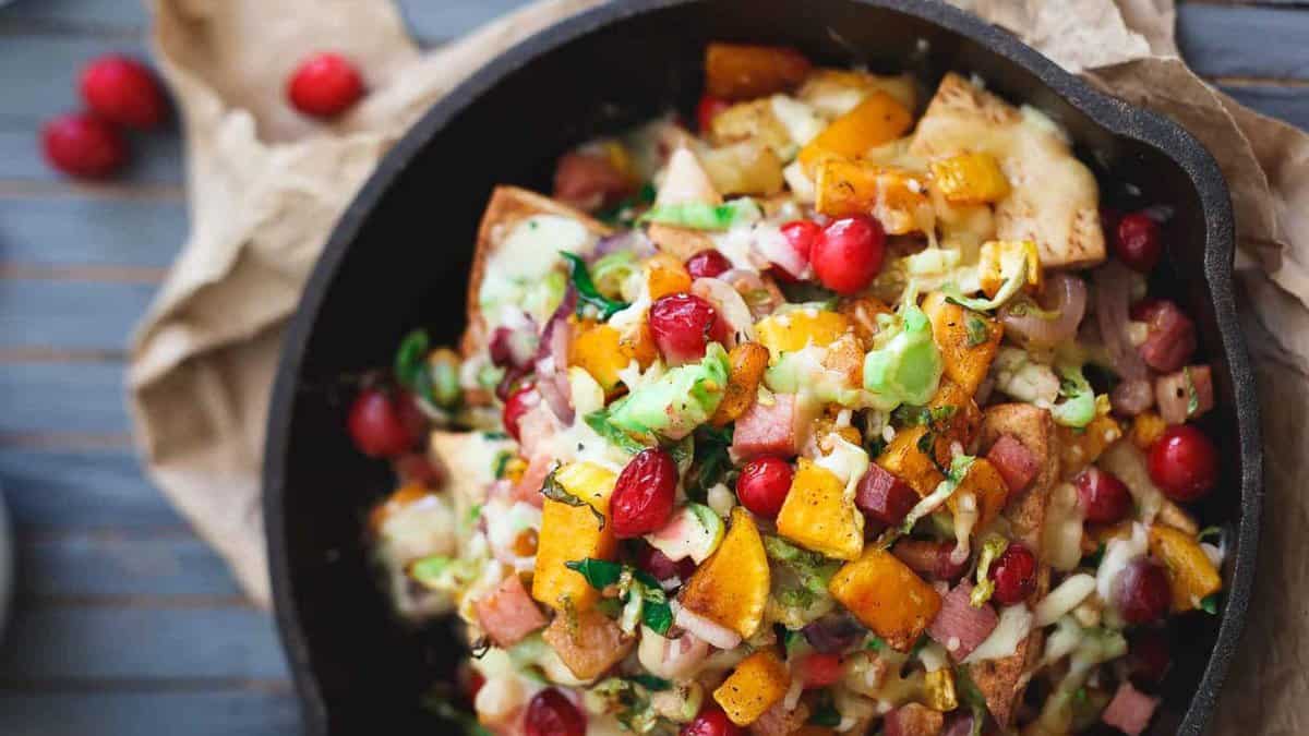 A cast iron skillet filled with roasted vegetables, diced ham, melted cheese, and whole cranberries on a wooden surface.