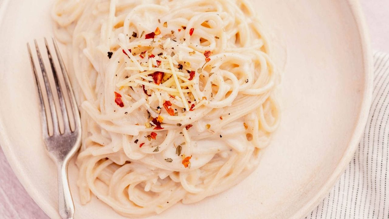 A plate of creamy spaghetti topped with shredded cheese, herbs, and red pepper flakes, with a fork placed on the side.