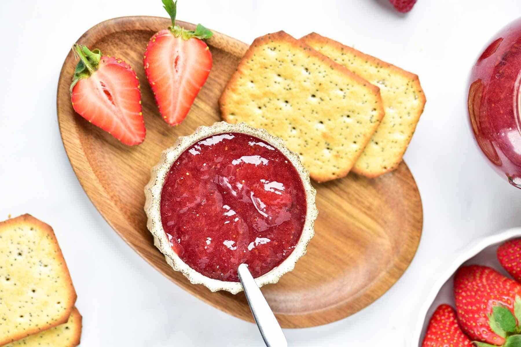 A wooden tray with strawberry jam in a bowl, a spoon, two halved strawberries, and three rectangular poppy seed crackers.