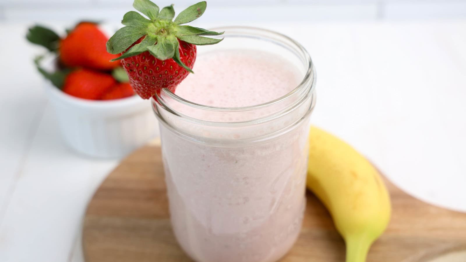 A glass jar filled with a pink smoothie, garnished with a strawberry, sits on a wooden board next to a banana and a bowl of strawberries.