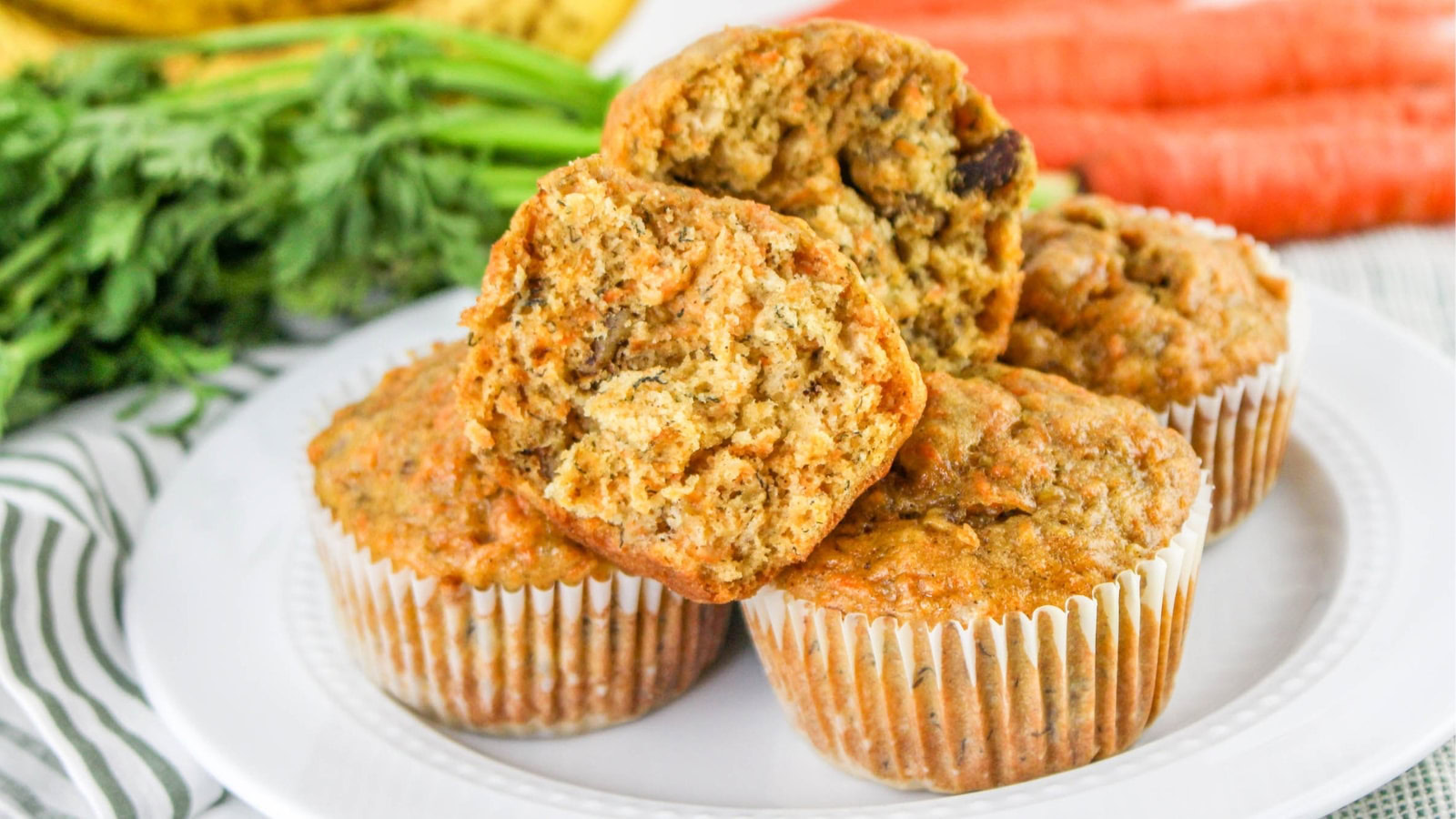 A plate of carrot muffins is shown, with one muffin cut in half to display the moist, textured interior. Fresh carrots and bananas are visible in the background.