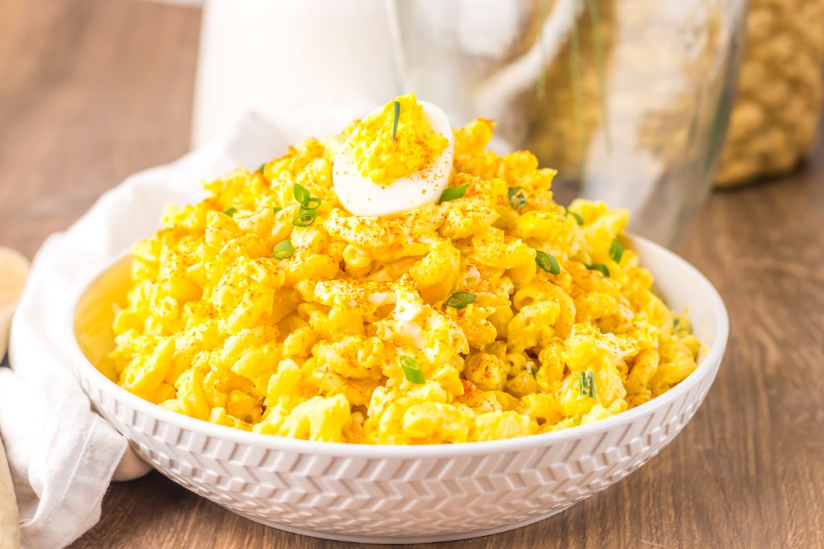 A bowl of creamy macaroni salad topped with sliced boiled eggs, green onions, and a sprinkle of paprika sits on a wooden table.