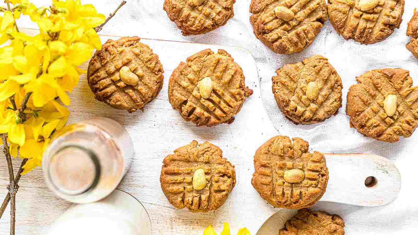 Peanut butter cookies with crisscross fork marks and peanuts on top are arranged on a white board next to a bottle of milk and yellow flowers.