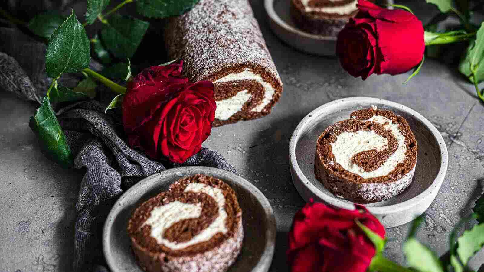 Chocolate Swiss roll cake with cream filling is sliced and served on plates, accompanied by red roses and a dark cloth on a gray surface.