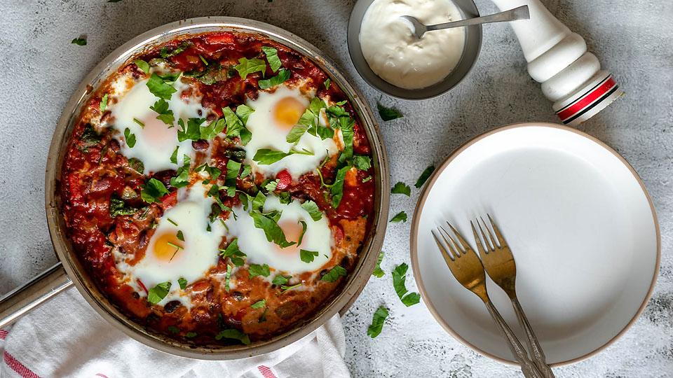 A skillet of shakshuka with eggs, tomato sauce, and herbs sits next to a small bowl of yogurt, an empty plate, and silverware on a light surface.