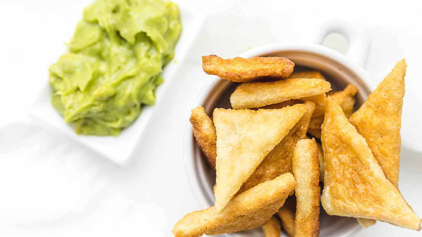 A cup filled with golden, crispy pita chips next to a small dish of smooth guacamole on a white background.
