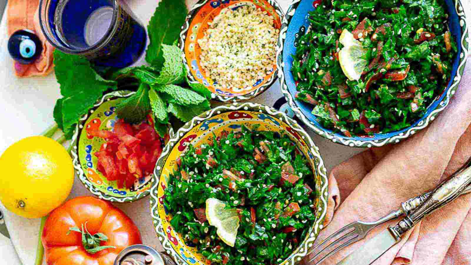 A colorful spread of tabbouleh salad garnished with lemon, chopped tomatoes, hemp seeds, fresh mint, lemon, a tomato, and a glass of water on a table.