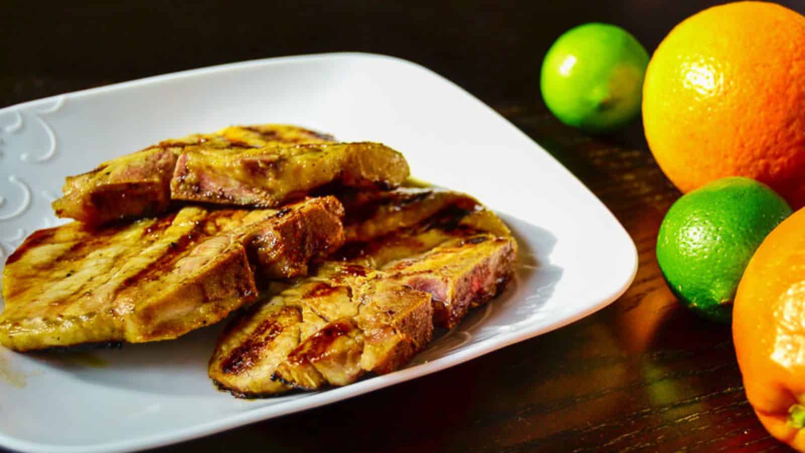 Grilled pork chops are stacked on a white plate, with an orange and two limes placed on the dark wooden table beside it.