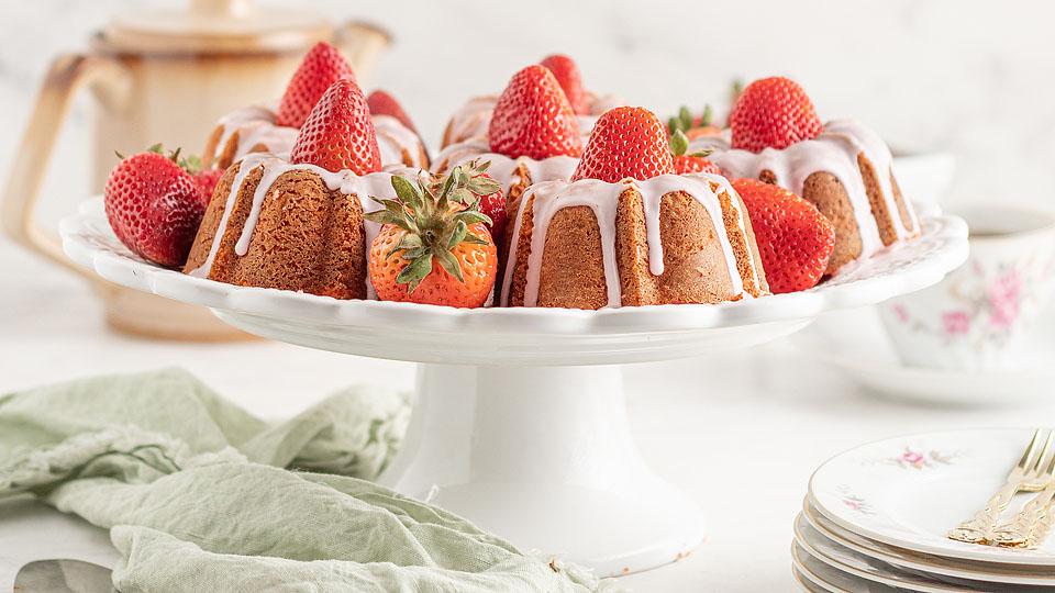 A bundt cake topped with whole strawberries and white icing sits on a white cake stand, with plates and a teapot in the background.