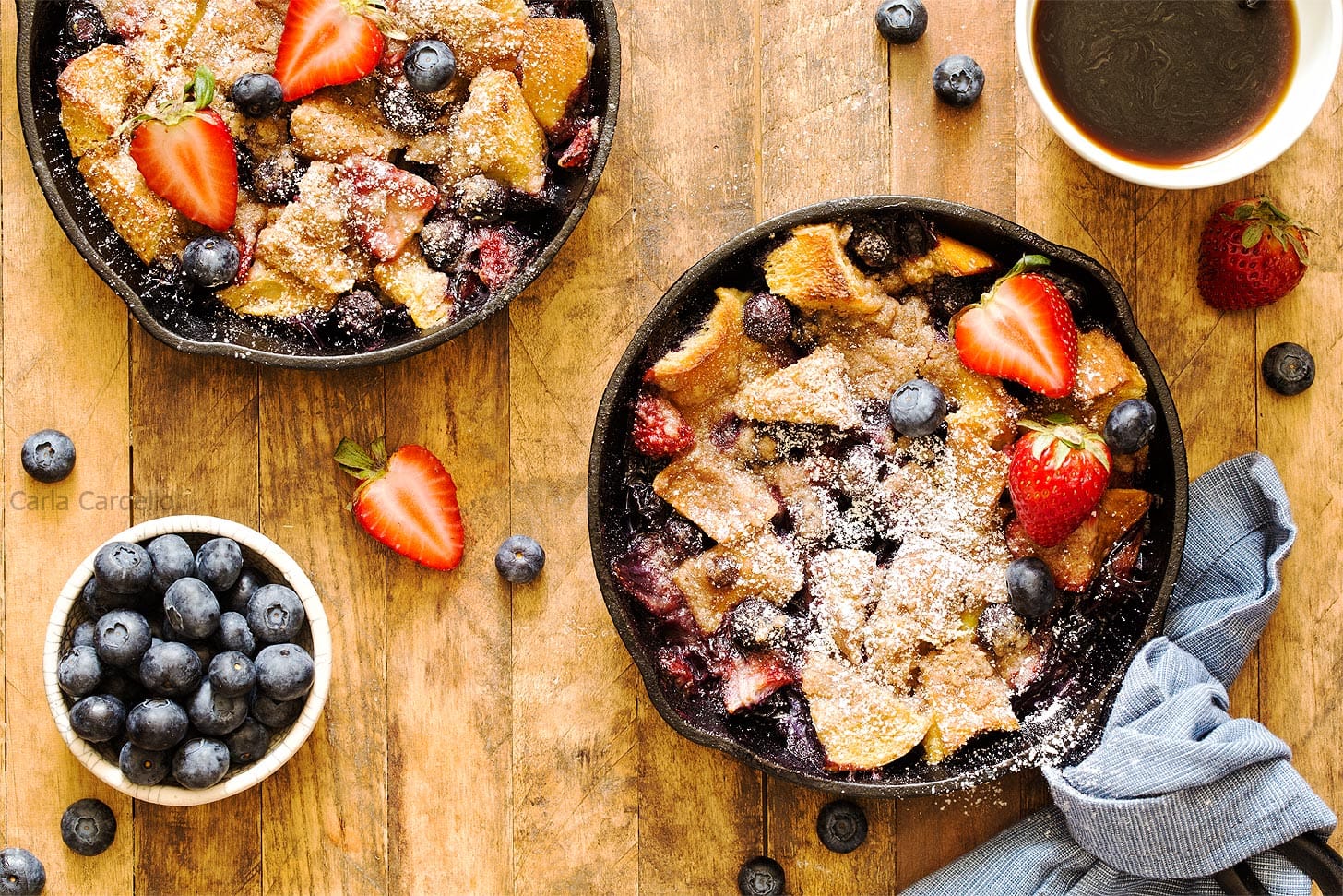 Two mini skillets of berry bread pudding topped with powdered sugar and fresh strawberries, surrounded by blueberries and a cup of coffee on a wooden table.