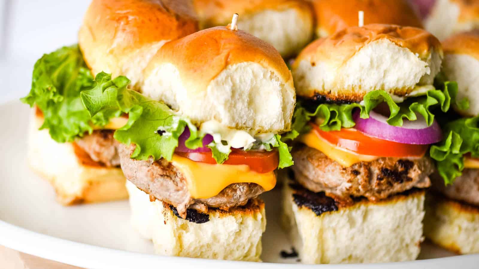 A plate of mini cheeseburgers on soft buns with lettuce, tomato, red onion, cheese, and burger patties.