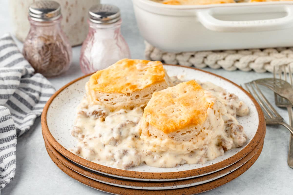 Two biscuits served on a plate with sausage gravy, with salt and pepper shakers and a baking dish in the background.