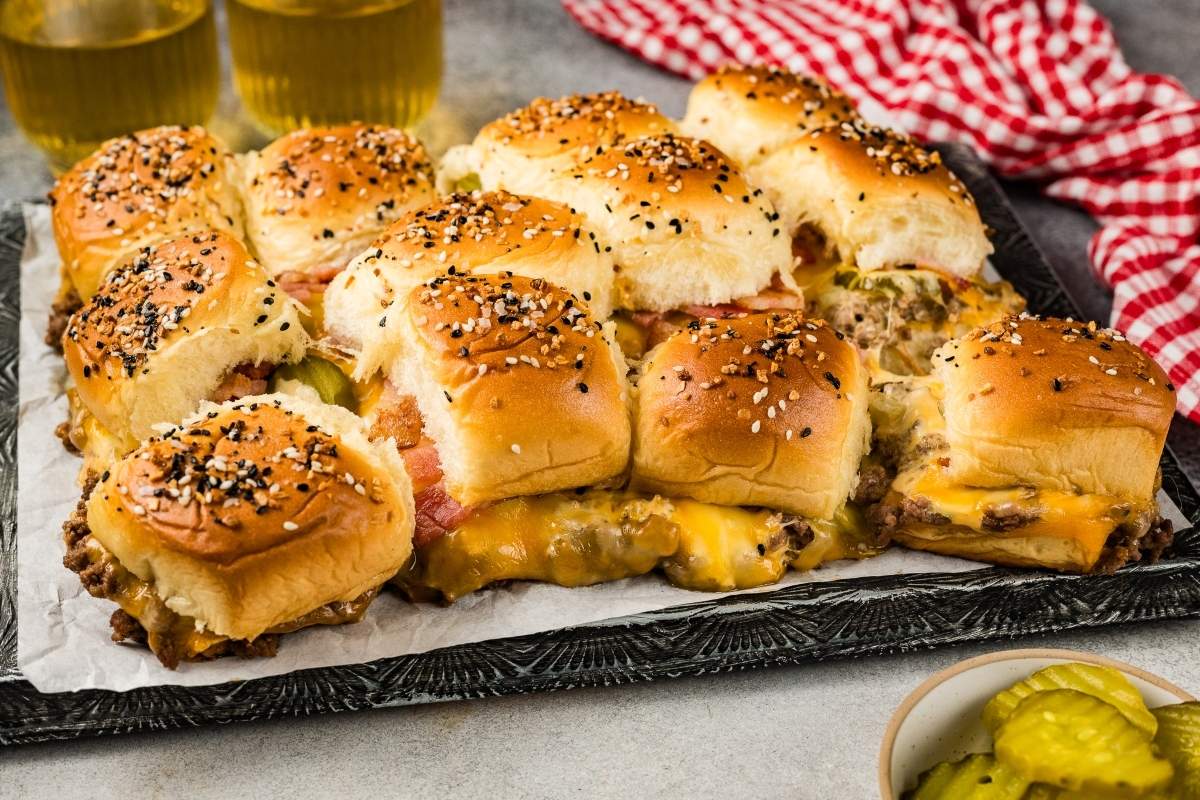 A tray of cheeseburger sliders with sesame seed-topped buns, melted cheese, pickles, and ground beef, with a red checked cloth and two glasses of drinks in the background.