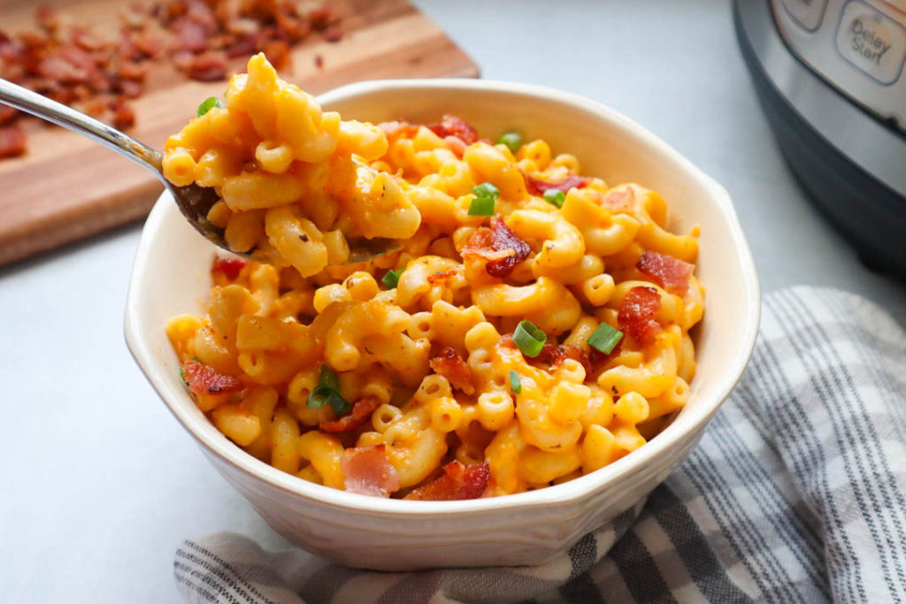A bowl of macaroni and cheese topped with bacon bits and chopped green onions, with a spoonful being lifted from the bowl.