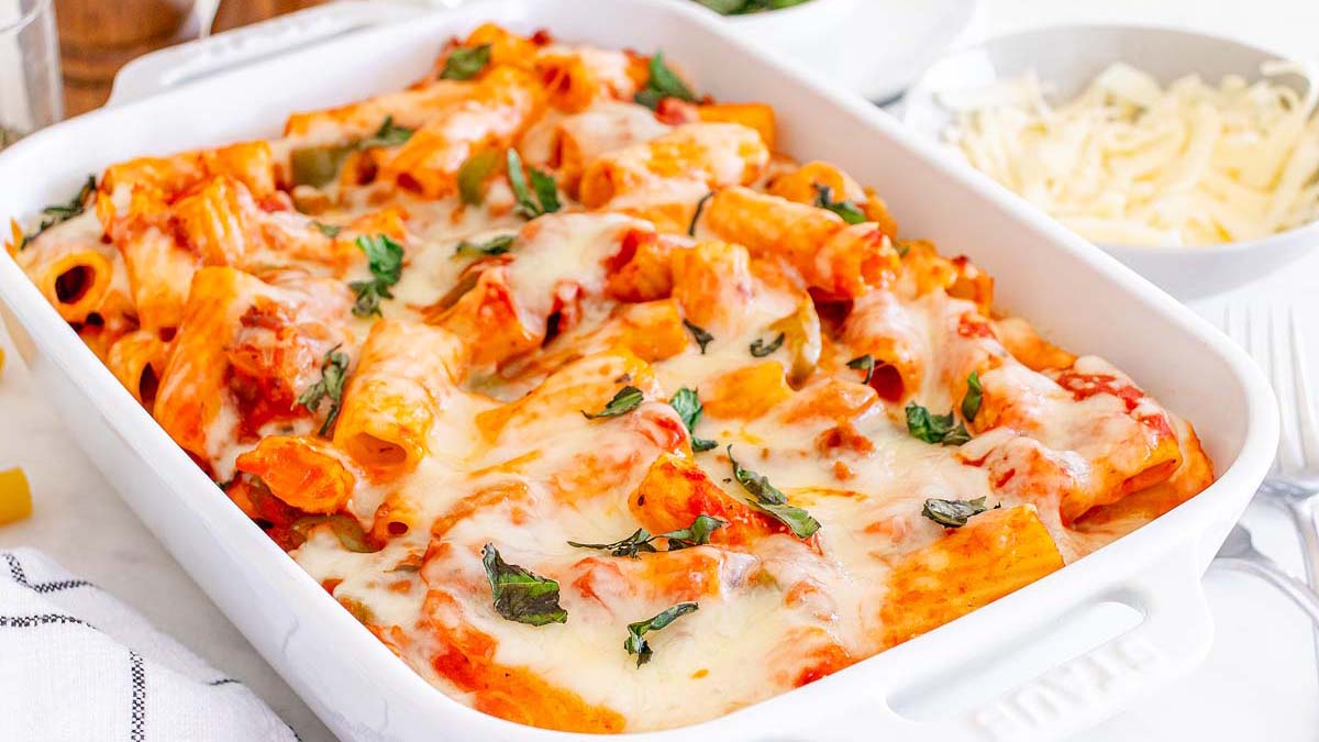 A white baking dish filled with baked rigatoni pasta in tomato sauce, topped with melted cheese and fresh basil.