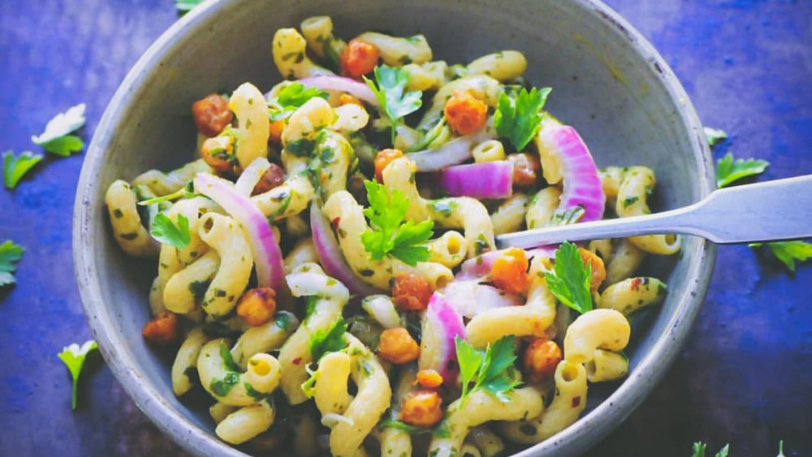 A bowl of macaroni pasta salad with chickpeas, red onion slices, chopped parsley, and a fork on the side.