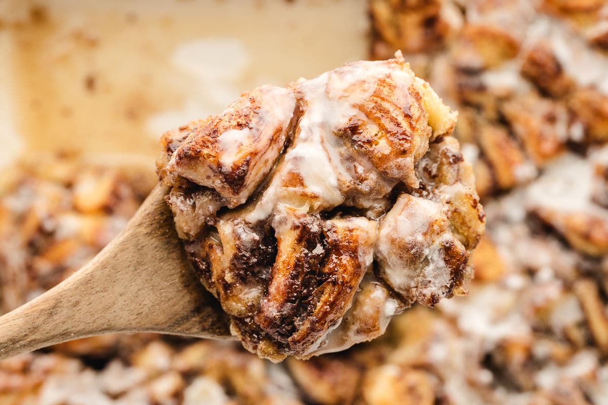 A wooden spoon holds a serving of cinnamon roll bread pudding topped with icing, with more bread pudding visible in the background.