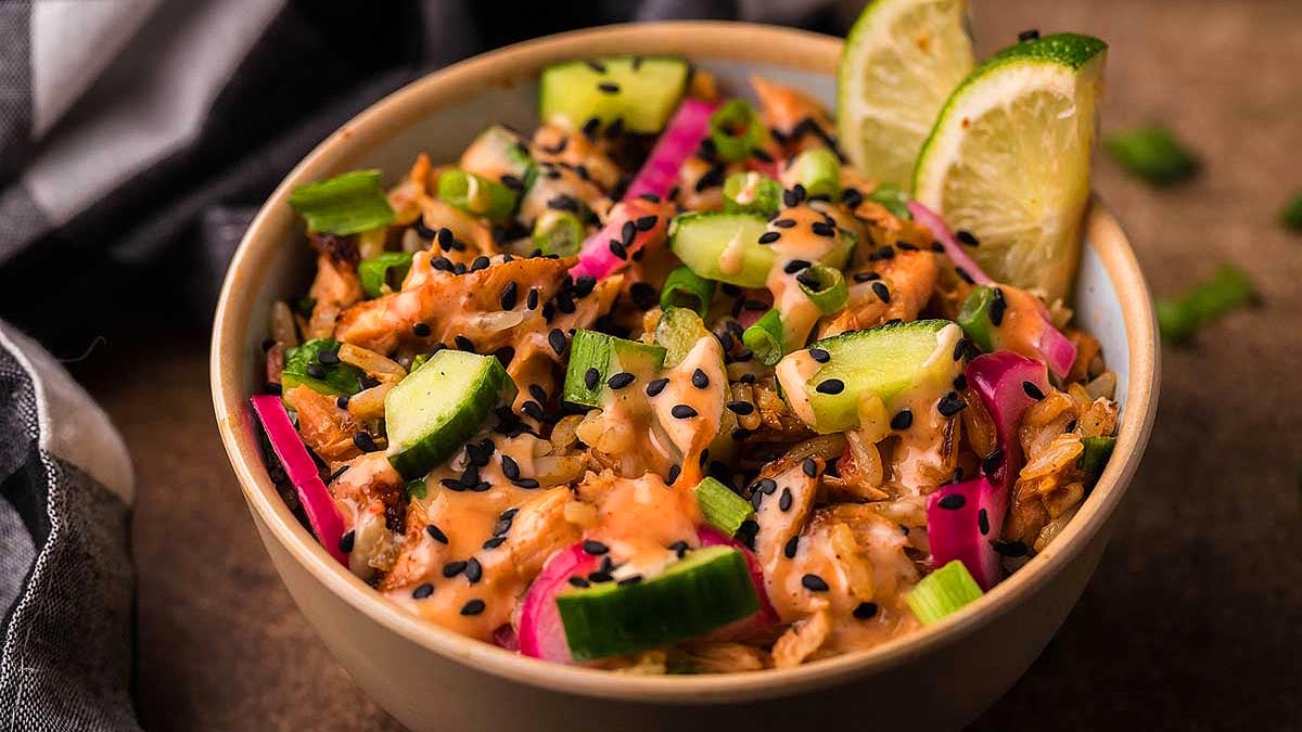 A bowl of rice topped with shredded chicken, sliced cucumbers, pickled onions, green onions, black sesame seeds, creamy sauce, and lime wedges.