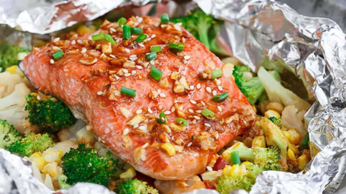 A cooked salmon fillet with sauce and chopped green onions sits on top of mixed vegetables, all wrapped in aluminum foil.