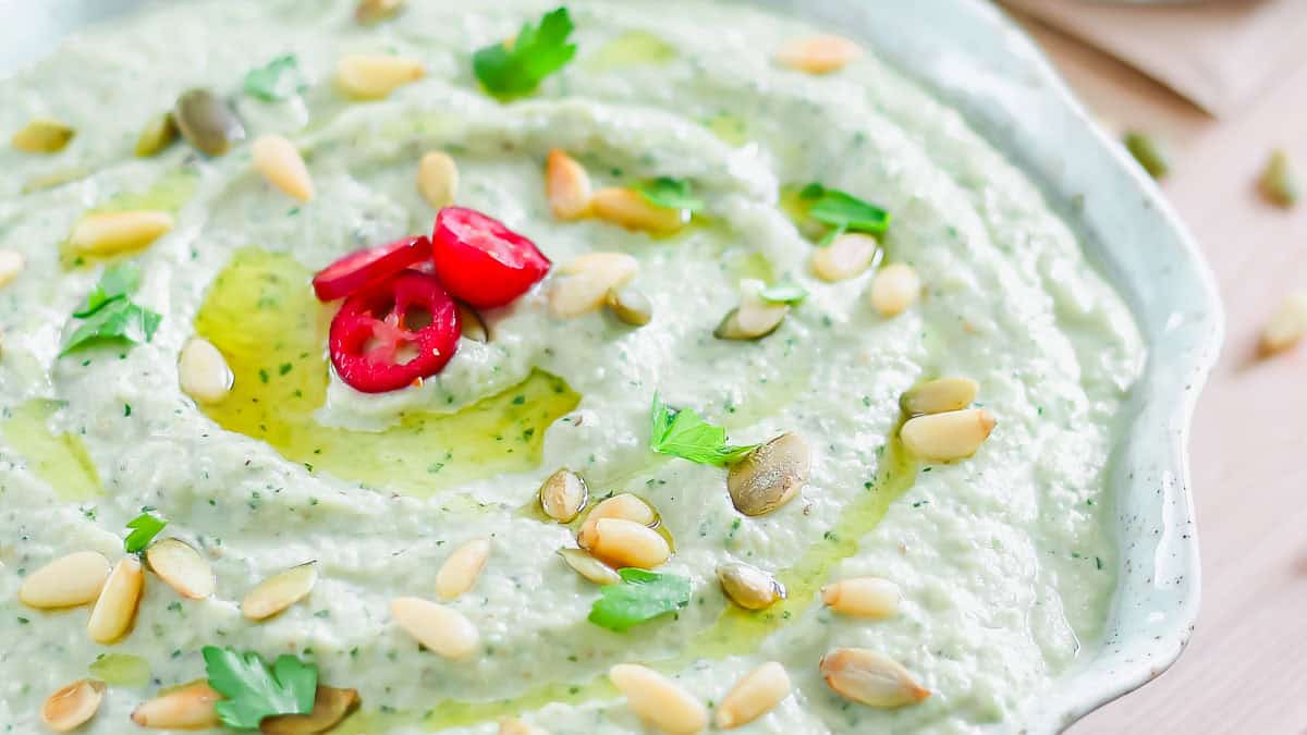 A plate of creamy dip garnished with sliced red peppers, pine nuts, pumpkin seeds, parsley, and a drizzle of olive oil.