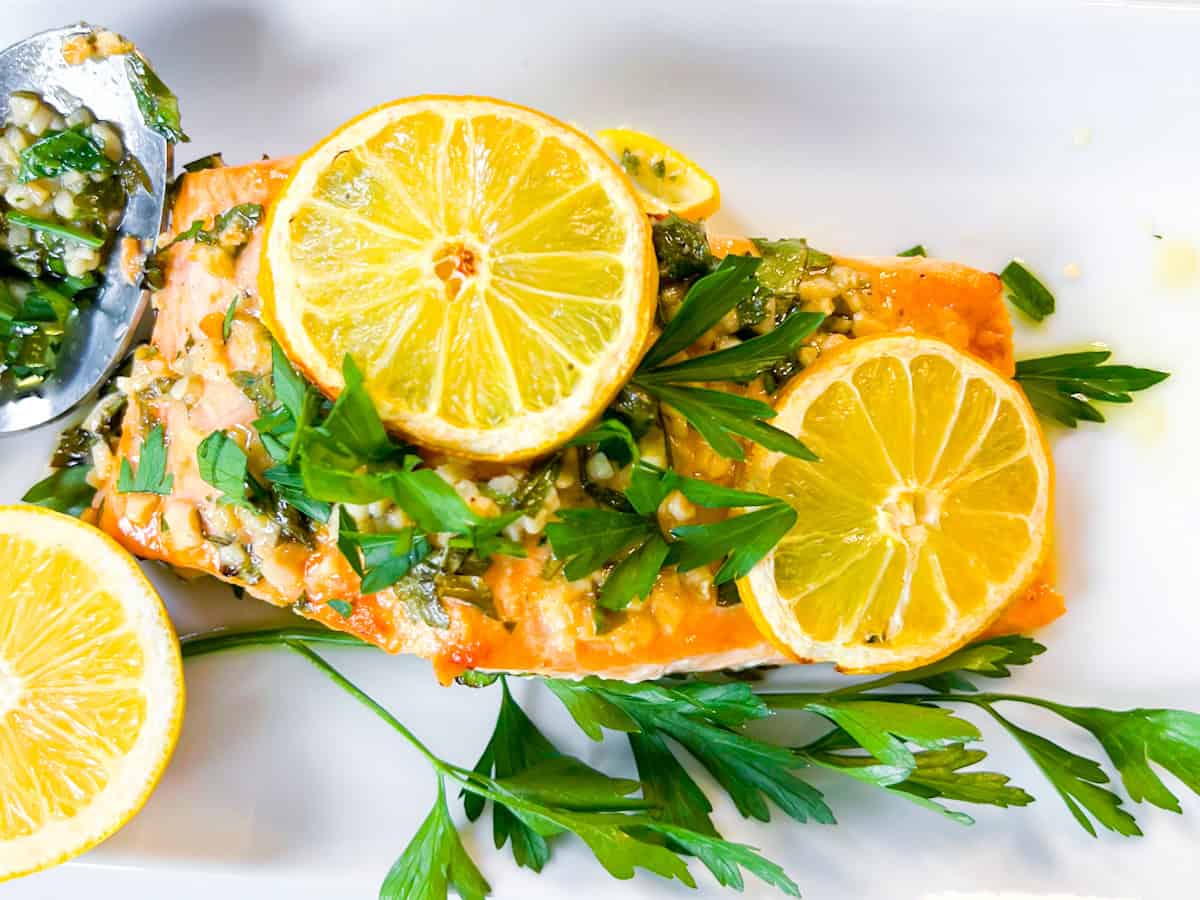 A cooked salmon fillet garnished with lemon slices, chopped herbs, and parsley on a white plate.