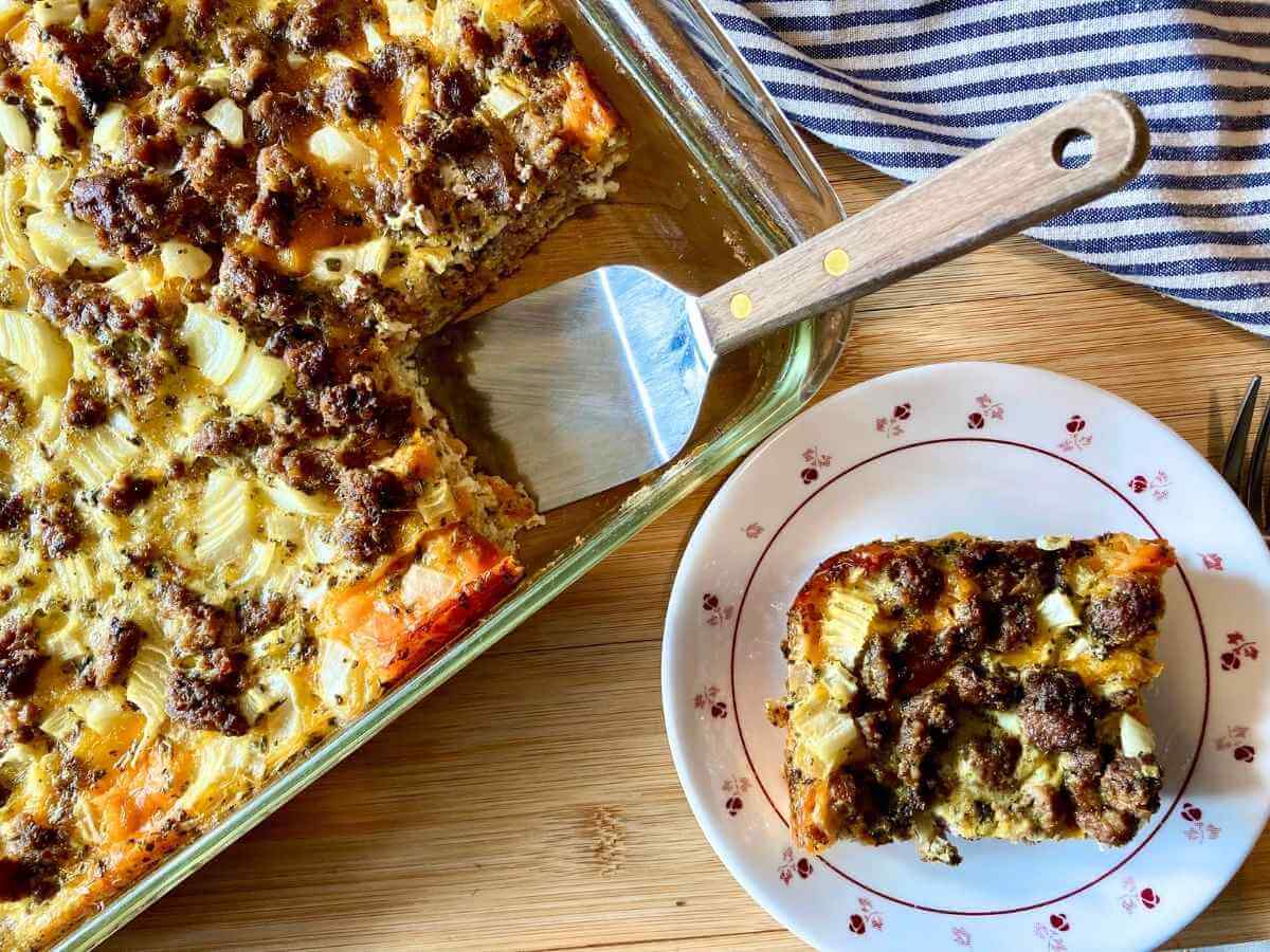 A slice of baked breakfast casserole with sausage and potatoes is served on a floral plate next to a glass baking dish and a serving spatula on a wooden surface.