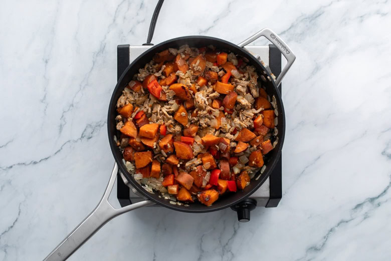 A skillet on a marble countertop contains a mixture of cooked rice, sweet potatoes, red bell peppers, and onions.