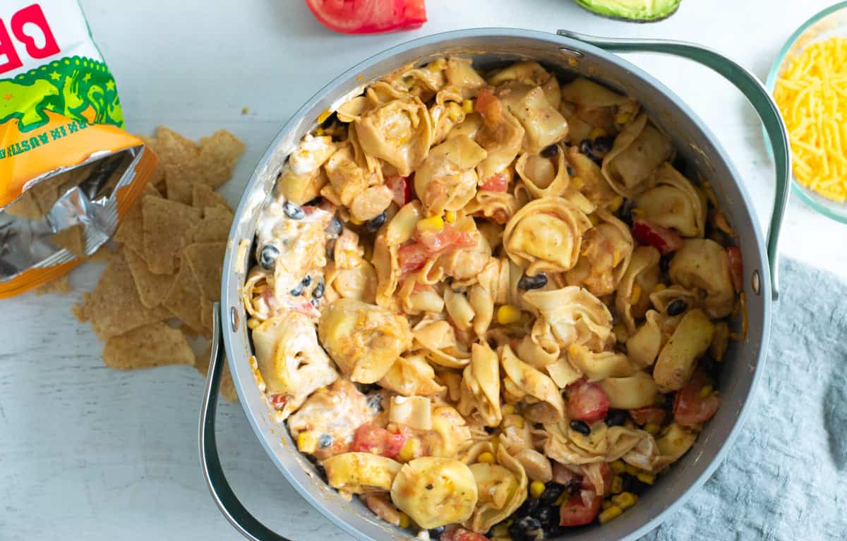 A pot filled with cheesy pasta, black beans, corn, tomatoes, and sauce sits on a white surface next to tortilla chips, a sliced tomato, avocado, and a bowl of shredded cheese.