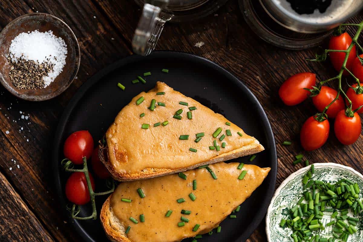 Two slices of toasted bread with melted cheese and chopped chives on a black plate, surrounded by cherry tomatoes, salt, pepper, and herbs on a wooden table.