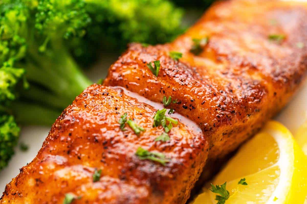 Close-up of a seasoned, cooked salmon fillet garnished with herbs, served with broccoli and lemon wedges.